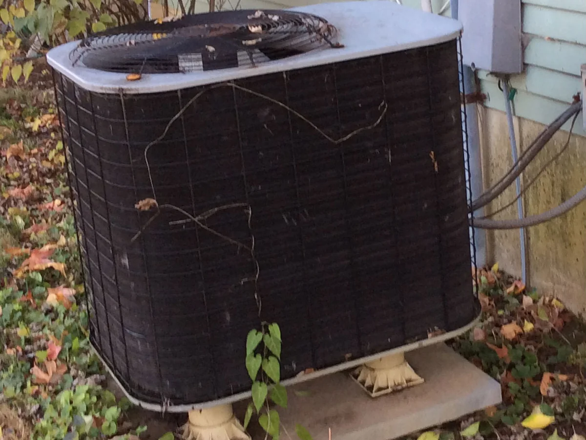 Old corroded air conditioning condenser with damaged fins and overgrown vegetation before replacement
