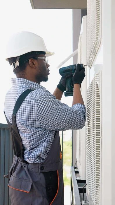 Altus Air technician in hard hat and safety glasses drilling into an outdoor air conditioning unit during a service call